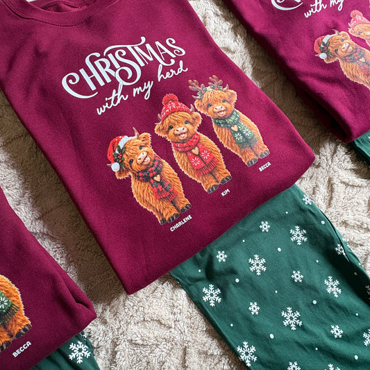 Highland Cow Family Christmas Loungewear set laid flat on a textured beige rug. The top is a burgundy sweatshirt featuring the white text 'CHRISTMAS with my herd' and three illustrated highland cows wearing Santa hats and scarves, with names printed below each bear.