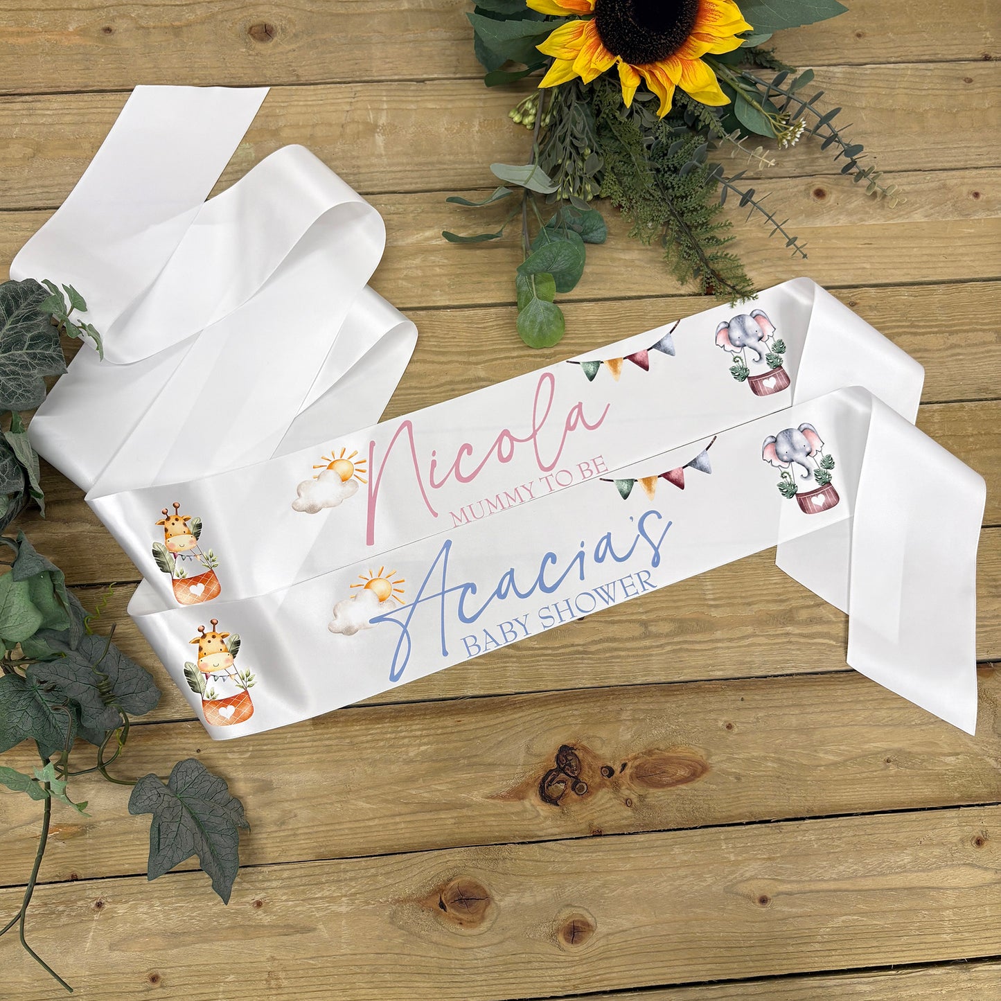 A white satin baby shower sash laid out on a wooden surface, in pink and blue script, decorated with small illustrations of safari animals and bunting. Sunflowers and greenery are styled around it.