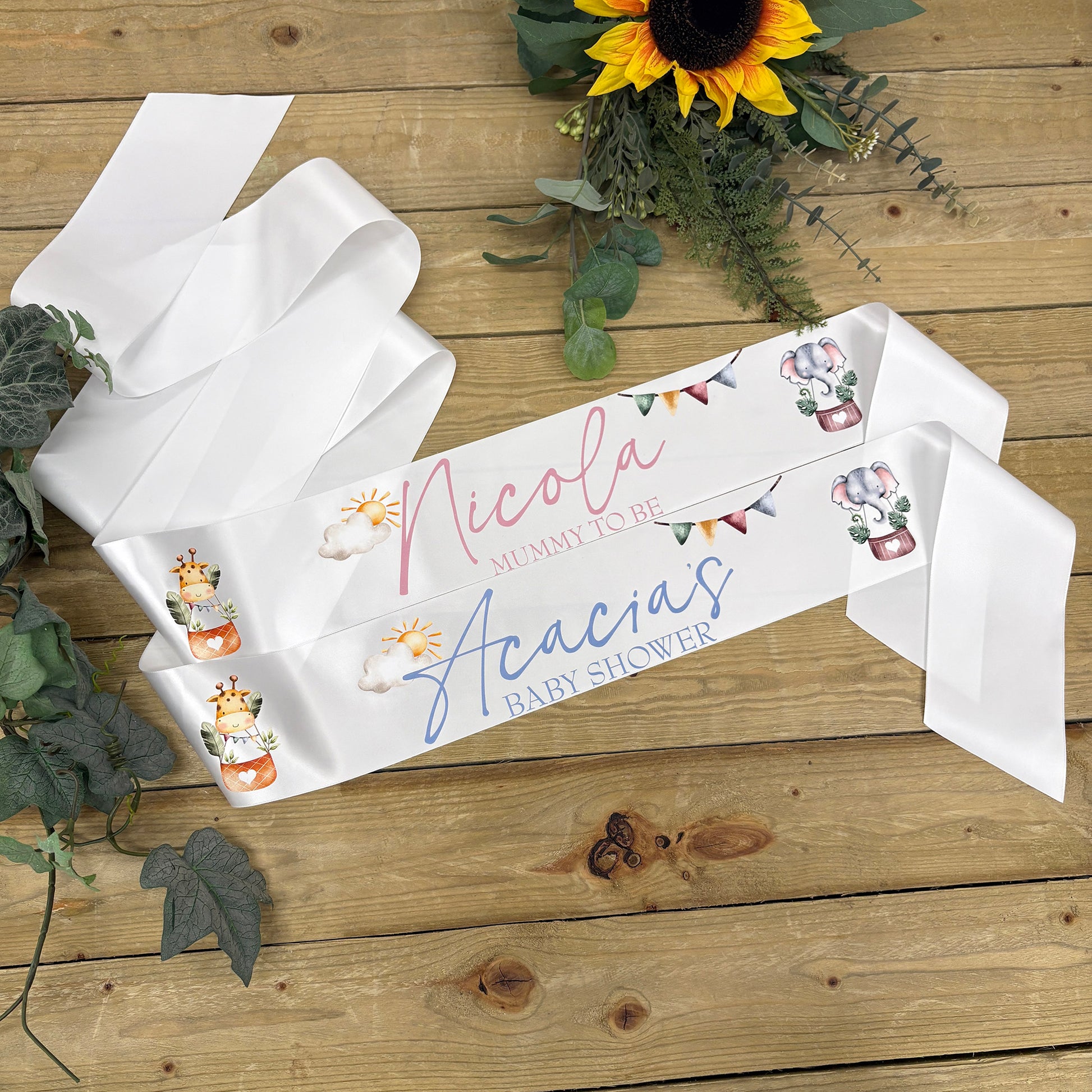 A white satin baby shower sash laid out on a wooden surface, in pink and blue script, decorated with small illustrations of safari animals and bunting. Sunflowers and greenery are styled around it.