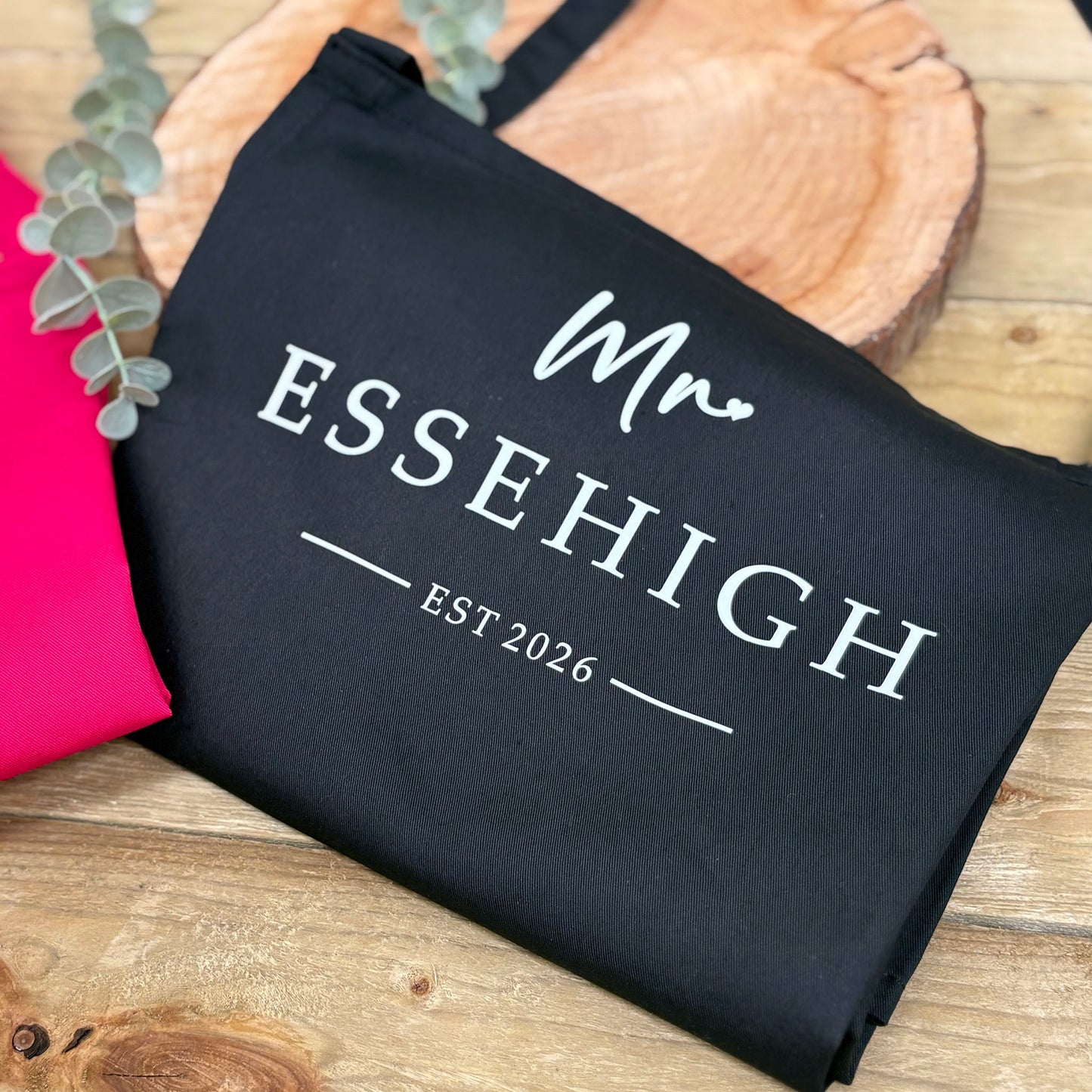 Black apron with 'Mr ESSEHIGH EST 2026' text on a wooden surface