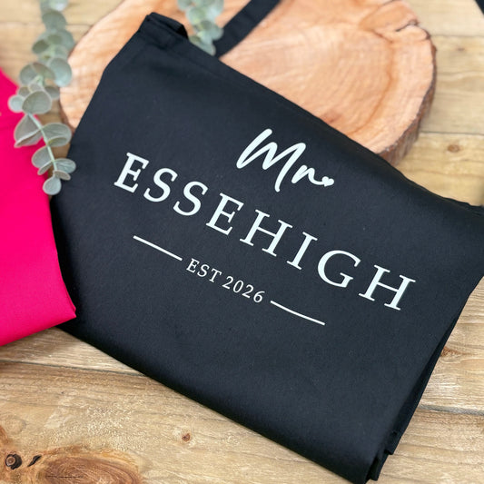 Black apron with 'Mr ESSEHIGH EST 2026' text on a wooden surface