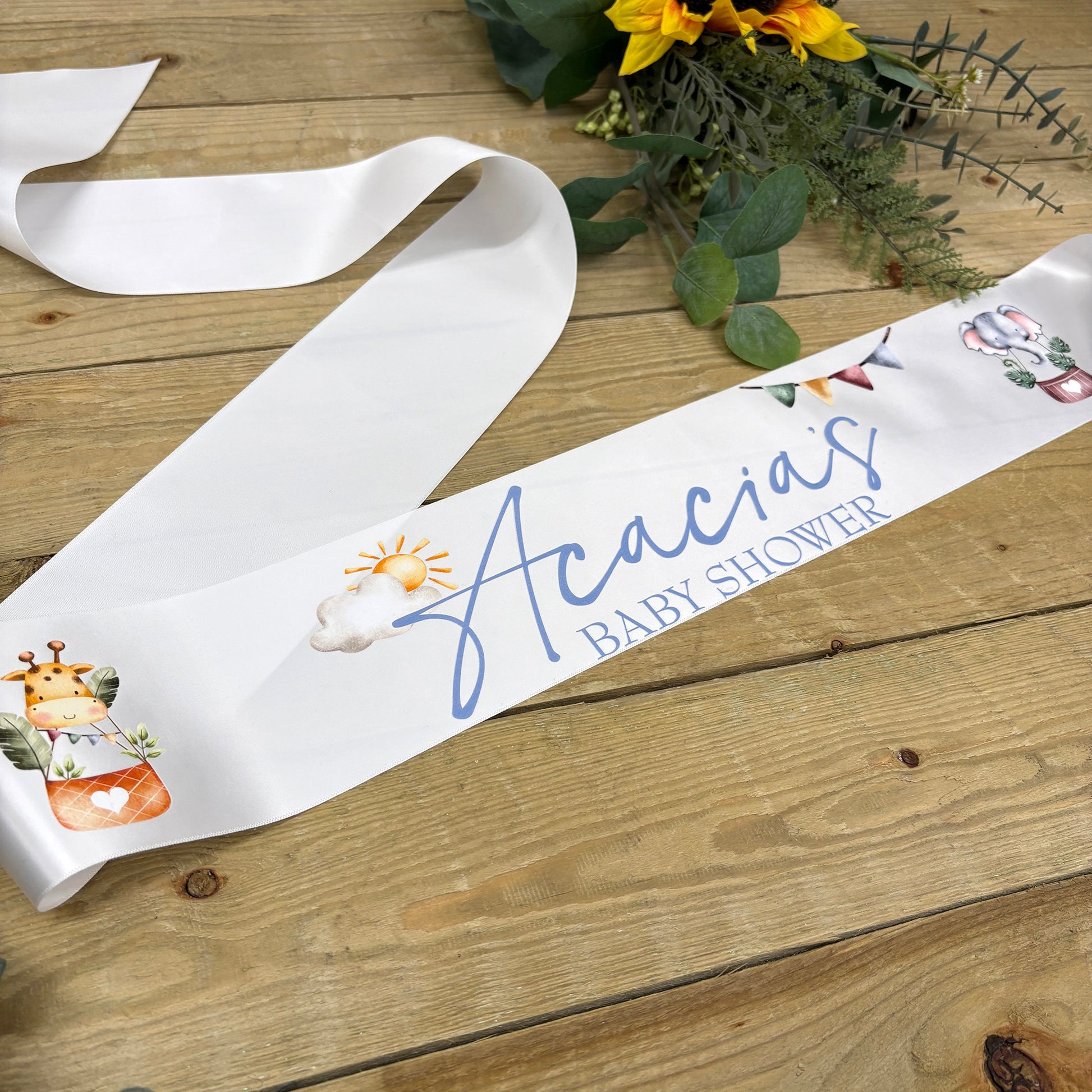 A white satin baby shower sash laid out on a rustic wooden surface, printed with the personalized name 'Acacia's Baby Shower' in blue script and decorated with small pastel illustrations. Yellow flowers and greenery are placed in the background as styling props.