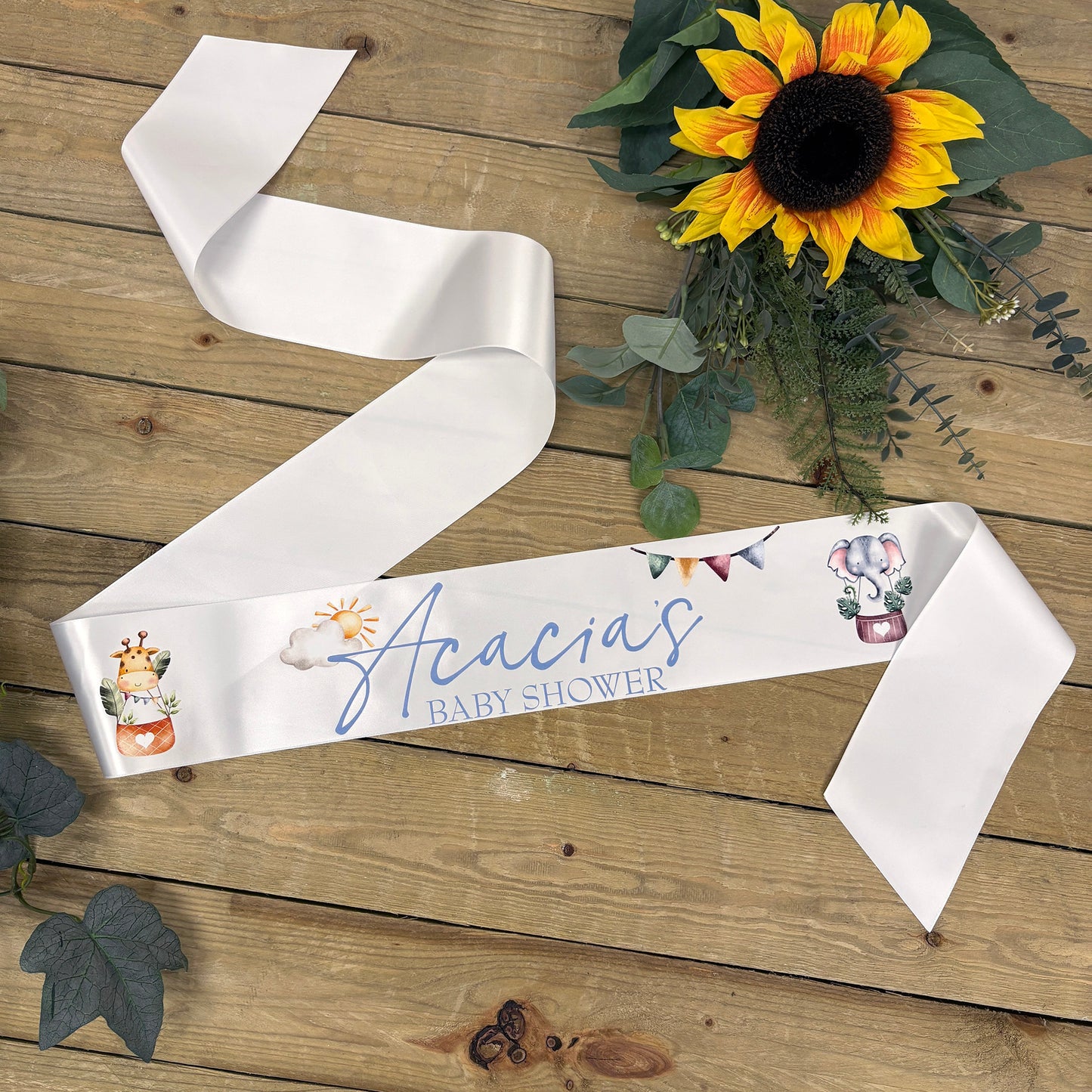 A white satin baby shower banner laid out on a rustic wooden surface, printed with the personalized text 'Acacia's BABY SHOWER' in blue script and decorated with small pastel illustrations. Sunflower and greenery props are placed around it.