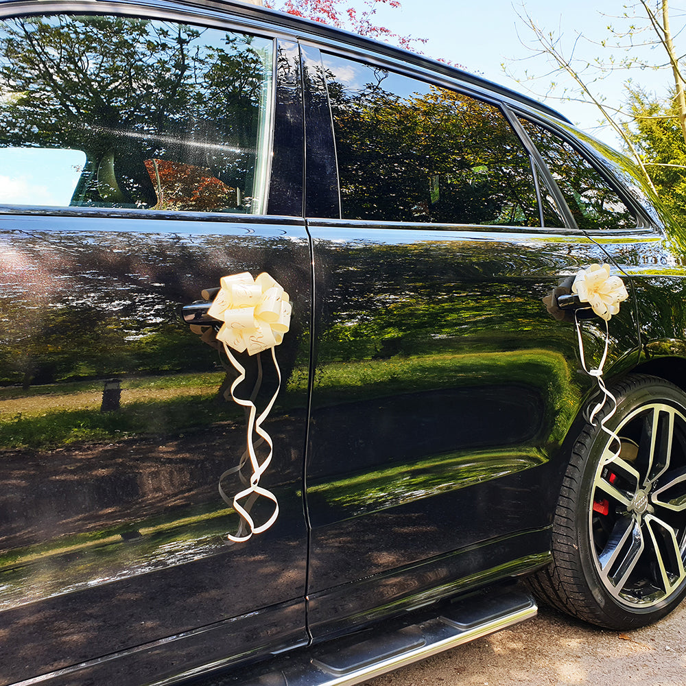 A black car with 2 ivory wedding car door bows attached.