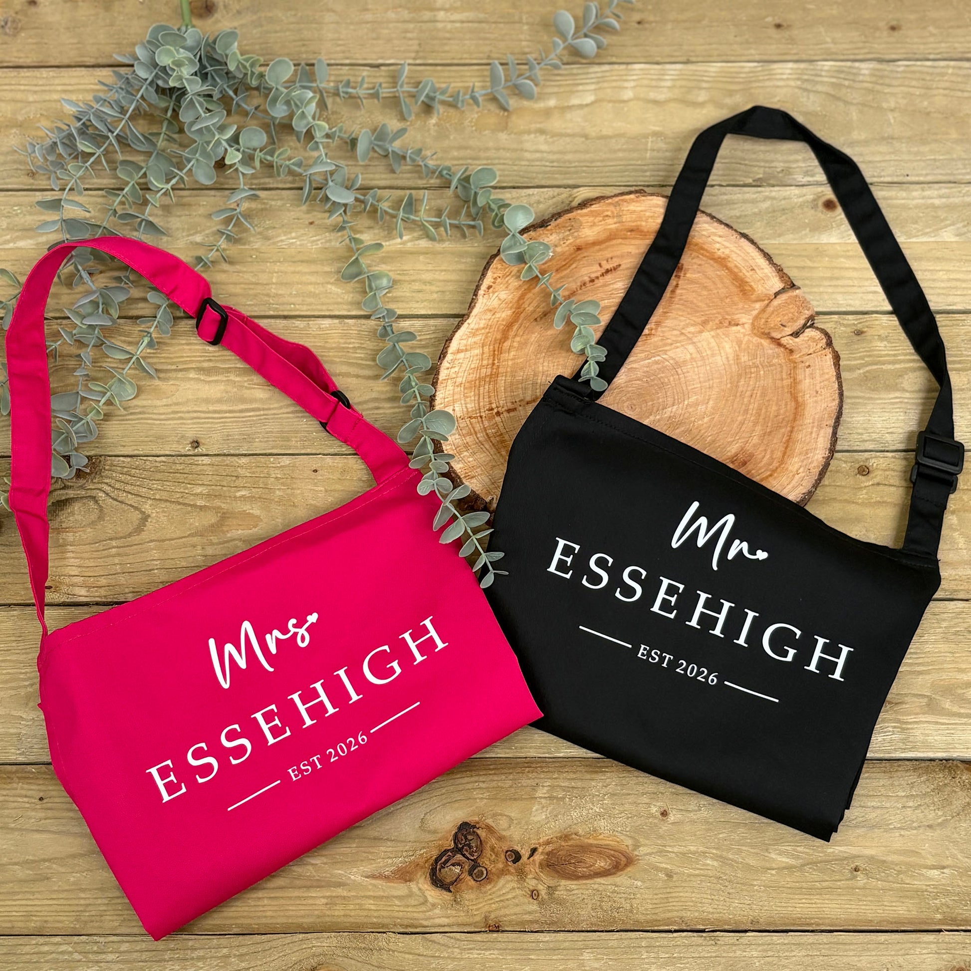 Pink and black aprons with 'Mrs ESSEHIGH' and 'Mr ESSEHIGH EST 2026' text on a wooden surface with eucalyptus leaves.