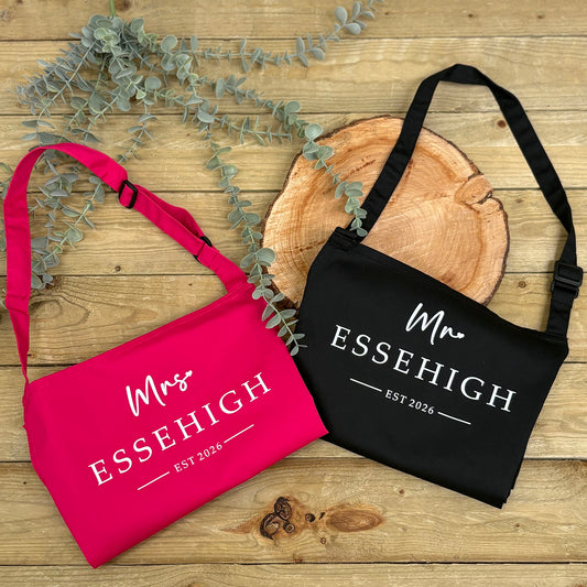 Pink and black aprons with 'Mrs ESSEHIGH' and 'Mr ESSEHIGH EST 2026' text on a wooden surface with eucalyptus leaves.