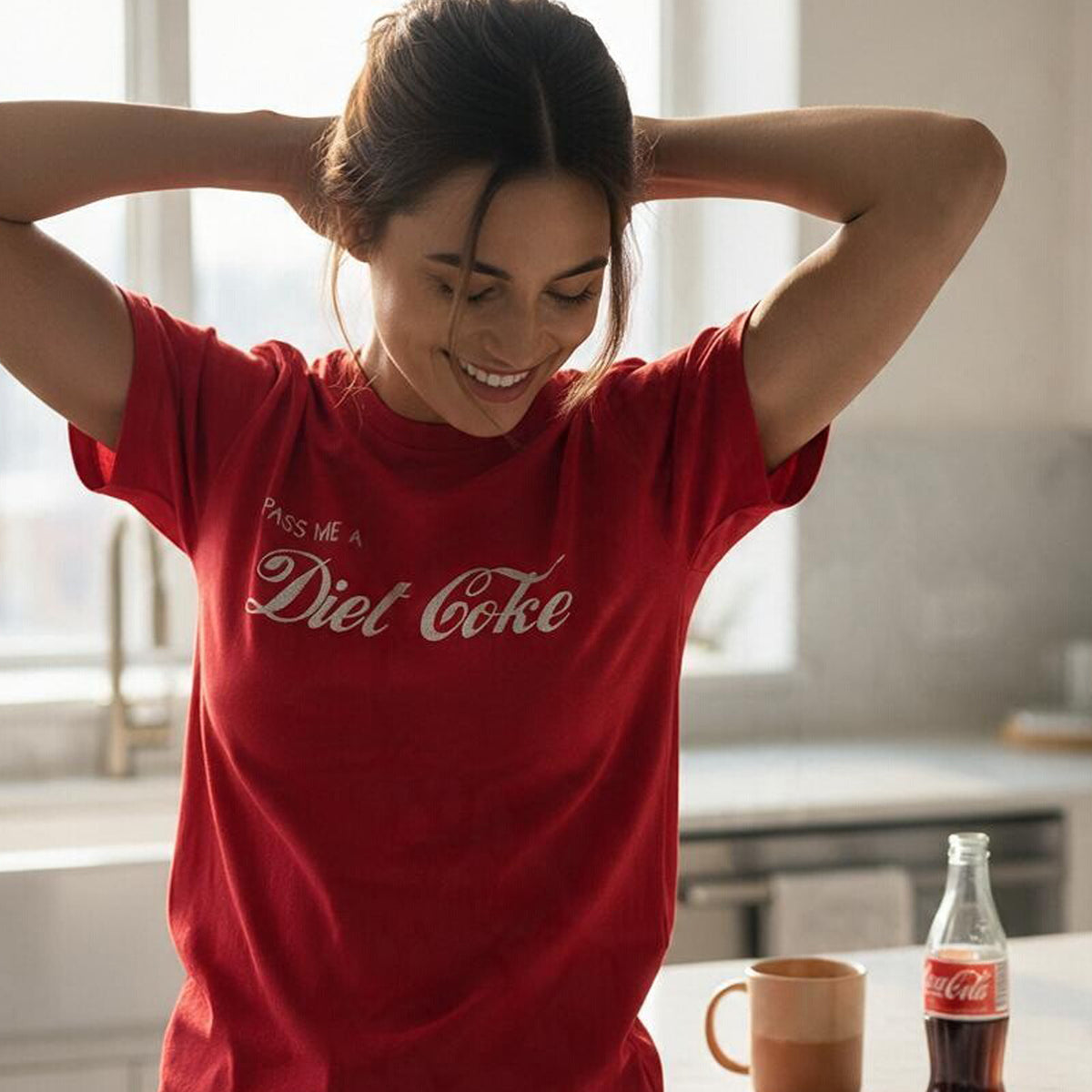 A woman in a kitchen wearing a red t-shirt with 'Pass Me A Diet Coke' printed in silver script. A glass Coca-Cola bottle and a mug sit on the counter in front of her.