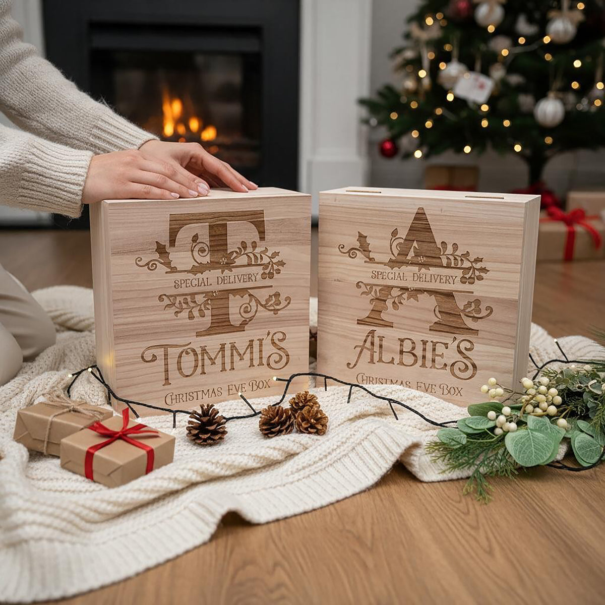 Two personalised wooden Christmas Eve boxes with engraved names and holiday designs, displayed on a cream knit blanket in a cosy living room setting with a fireplace, Christmas tree, and small wrapped gifts.