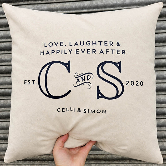 A square cream-colored cushion with personalised black text and monogram lettering, held by a hand against a grey corrugated metal background.