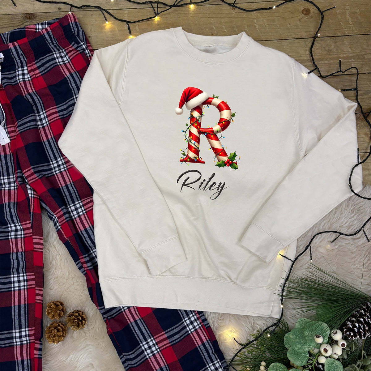 A cream crewneck sweatshirt laid flat on a rustic wood surface, featuring a large personalized letter 'R' Christmas design with candy canes, holly, and a Santa hat, with the name 'Riley' printed below. Red and navy tartan pyjama bottoms, fairy lights, pine needles, and small pinecones are styled around it.