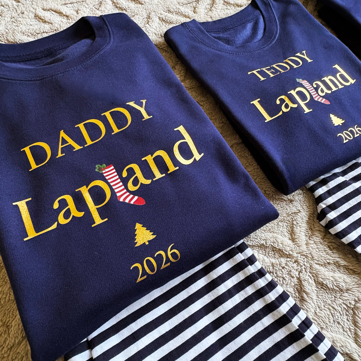 Personalised Navy Lapland Christmas Loungewear sets. Featuring Metallic Gold Print and a Red and White Striped stocking with Holly, a Gold Christmas tree and the year. Paired with Navy and White Striped Trousers.