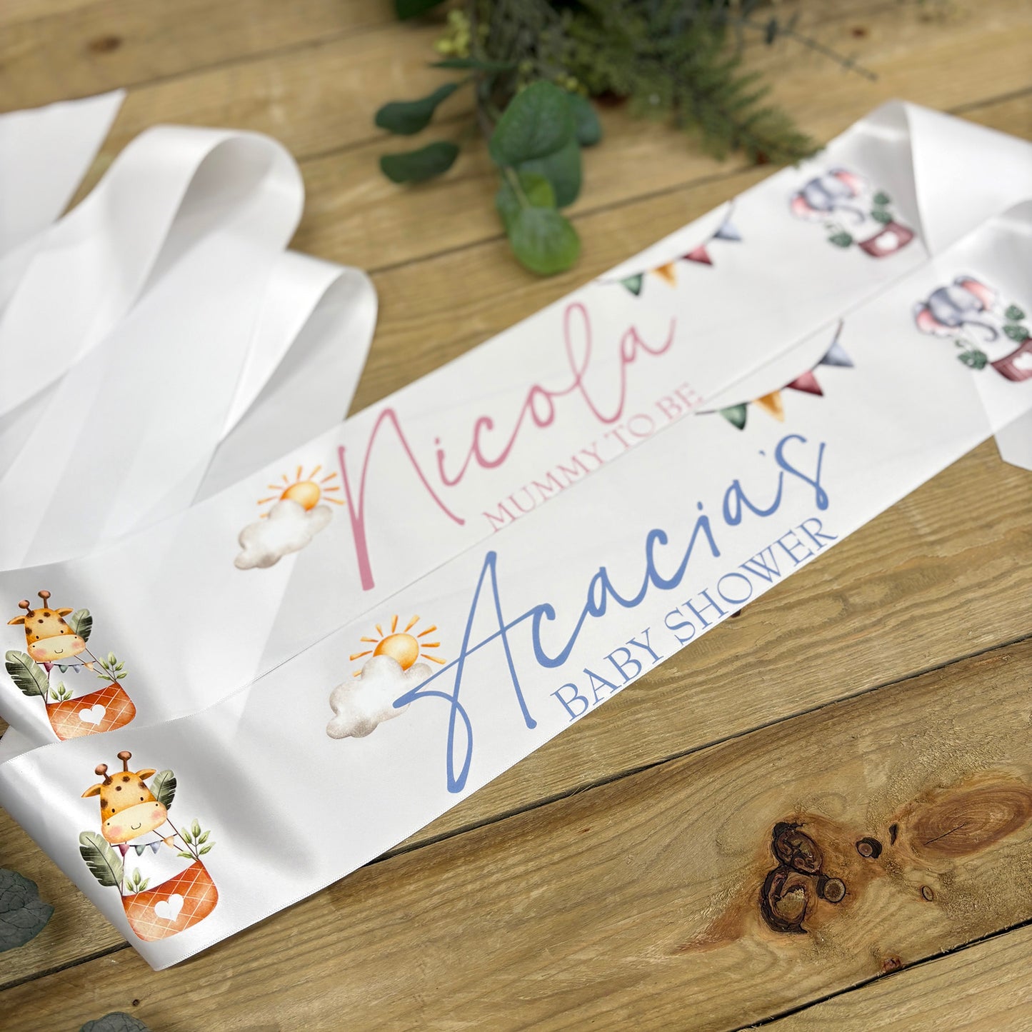 A white personalized baby shower sash laid diagonally on a rustic wooden surface, featuring custom printed text reading with pastel safari illustrations and bunting.