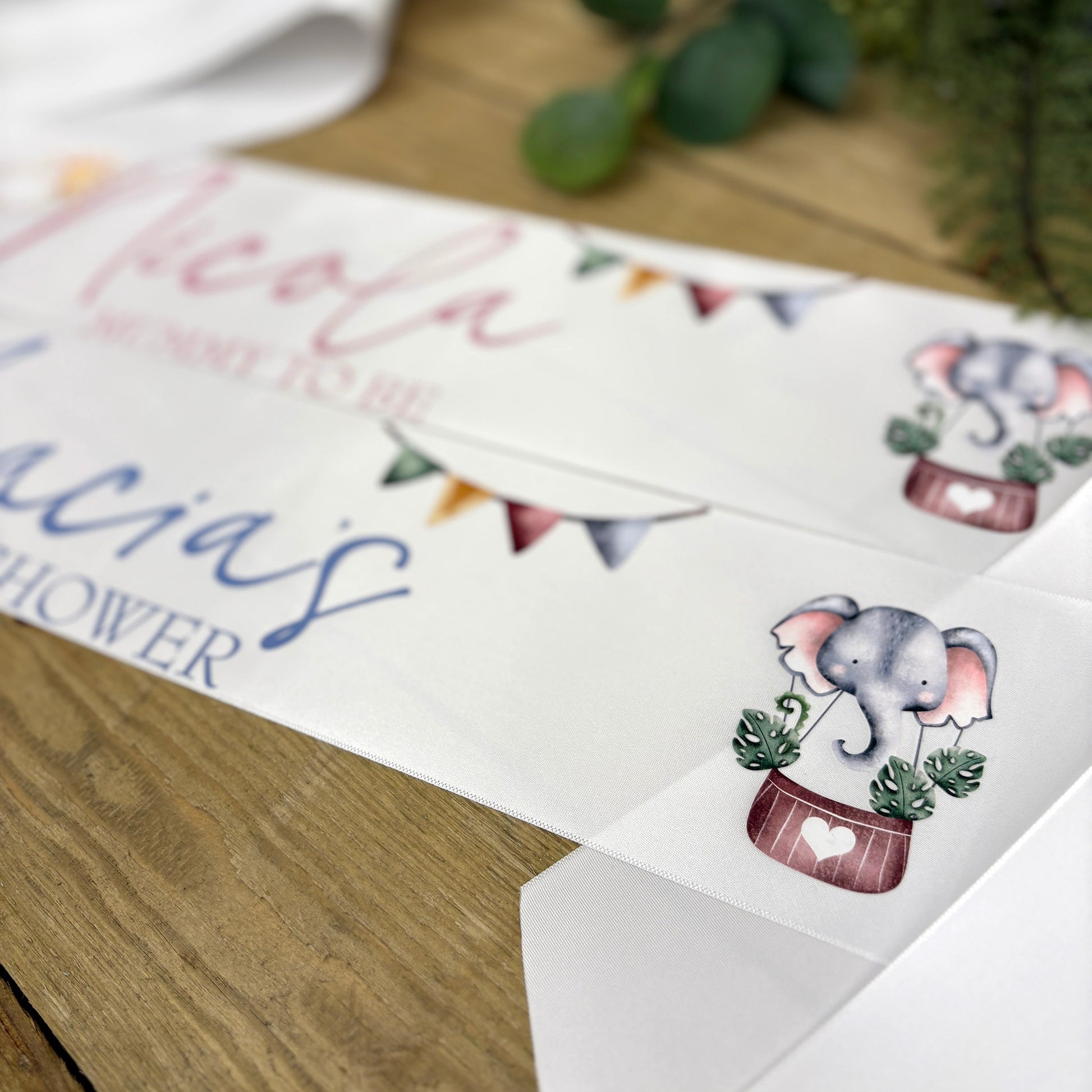 A close-up of a printed baby shower sashes laid on a wooden surface. The white sashes feature personalised text in pink and blue script, bunting flags, and illustrated elephants in planters with small green leaves.
