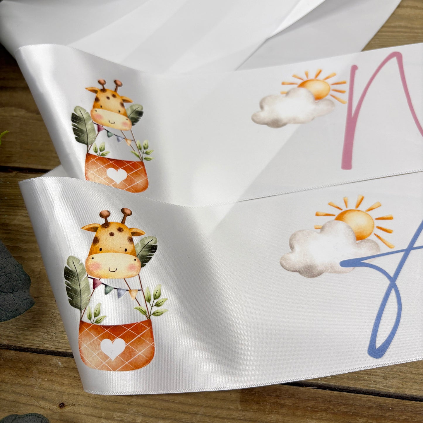 White satin baby shower sashes with cute cartoon giraffe and sun-and-cloud illustrations, displayed on a rustic wooden surface.
