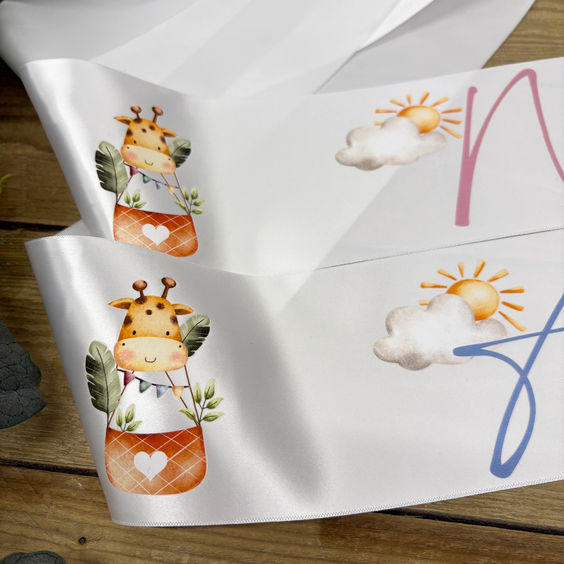 White satin baby shower sashes with cute cartoon giraffe and sun-and-cloud illustrations, displayed on a rustic wooden surface.