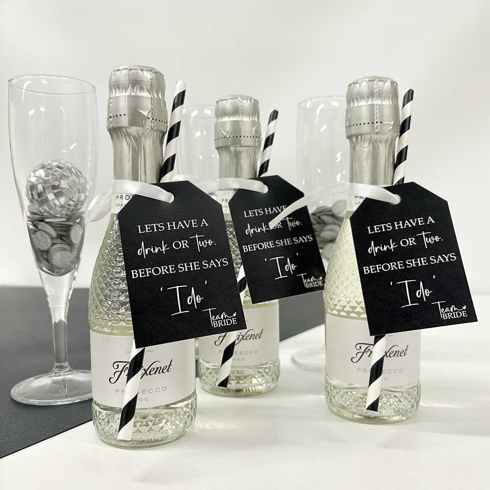 Three mini prosecco bottles with Team Bride Hen Party Bottle Tags attached. Printed with 'Let's have a drink or two before she says I do' text on black and white tags attached to them, displayed with black and white straws and a glass of champagne in the background.