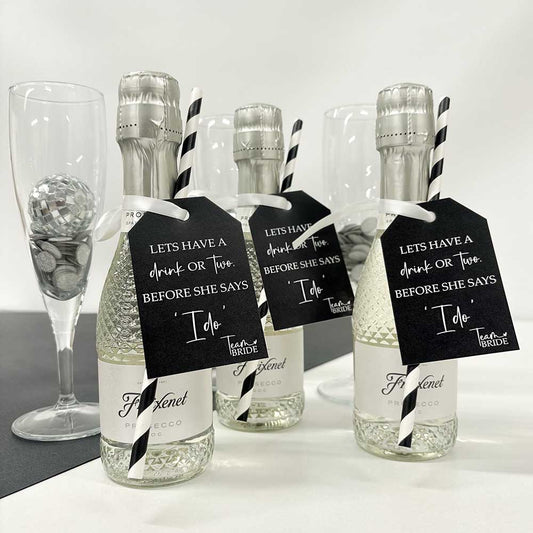 Three mini prosecco bottles with Team Bride Hen Party Bottle Tags attached. Printed with 'Let's have a drink or two before she says I do' text on black and white tags attached to them, displayed with black and white straws and a glass of champagne in the background.