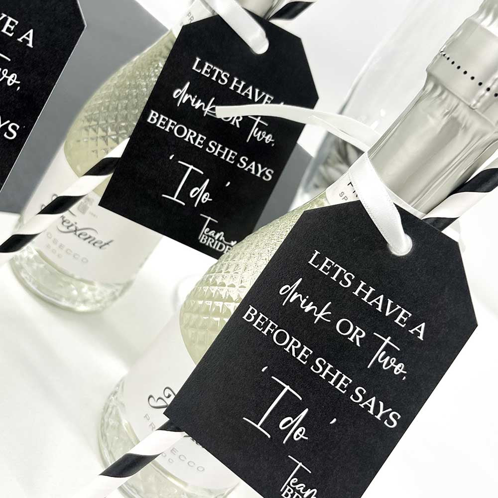 Two mini prosecco bottles with 'Team Bride Hen Party Bottle Tags attached. Printed with 'Let's have a drink or two before she says I do' text on black and white tags, displayed with black and white straws and a glass of champagne in the background.