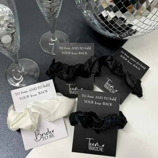 A collection of hen party scrunchies in black and white, with cards attached that read 'To have and to hold your hair back', displayed on a table with glasses and a mirror ball in the background.