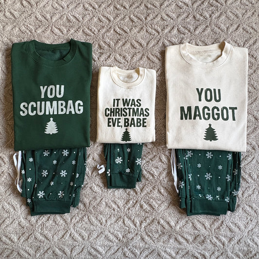 You Scumbag, You Maggot Family Christmas Loungewear. Three folded Christmas-themed sweatshirt and pyjama sets arranged side by side on a textured beige rug. The left set is dark green with the text 'YOU SCUMBAG' and a vanilla tree graphic; the middle set is vanilla with the text 'IT WAS CHRISTMAS EVE, BABE' and a dark tree graphic; the right set is vanilla with the text 'YOU MAGGOT' and a dark tree graphic.