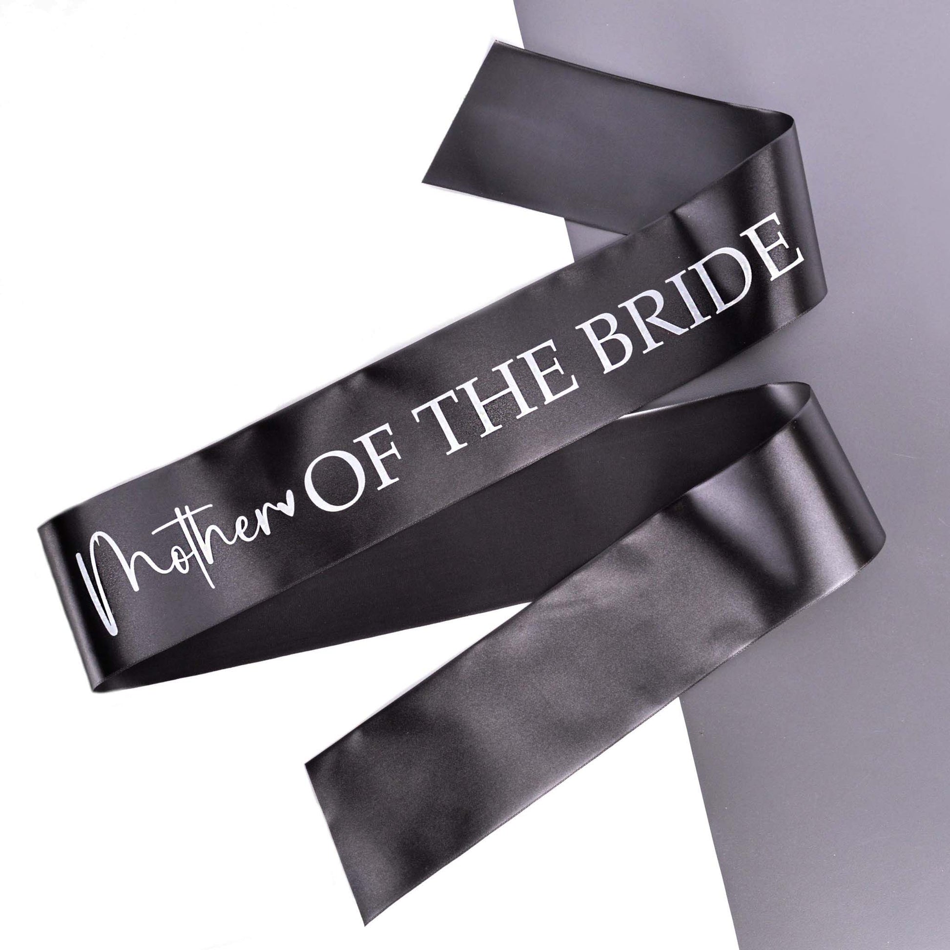 A Mother of the Bride Hern Party Sash printed in white on black ribbon, laid out on a grey background.