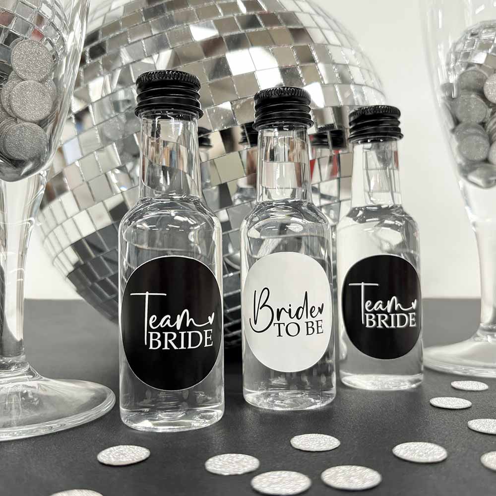 Three team bride hen party shot bottles with black screw caps, two labelled 'Team Bride' and one labelled 'Bride to Be', displayed on a table.