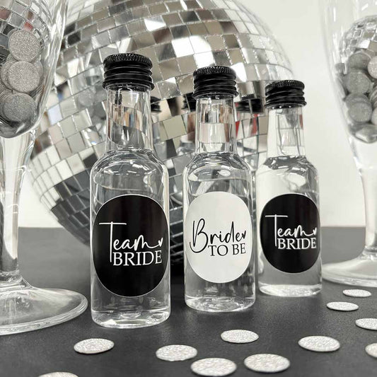 Three team bride hen party shot bottles with black screw caps, two labelled 'Team Bride' and one labelled 'Bride to Be', displayed on a table.